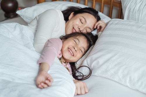 Image of mom and daughter laying down in bed, smiling, with white pillows and sheets