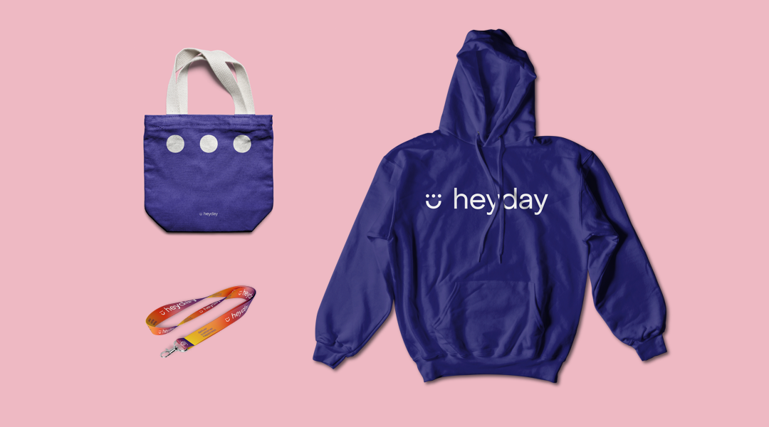 A tote bag, lanyard and hoodie on a pink background with Heyday logo