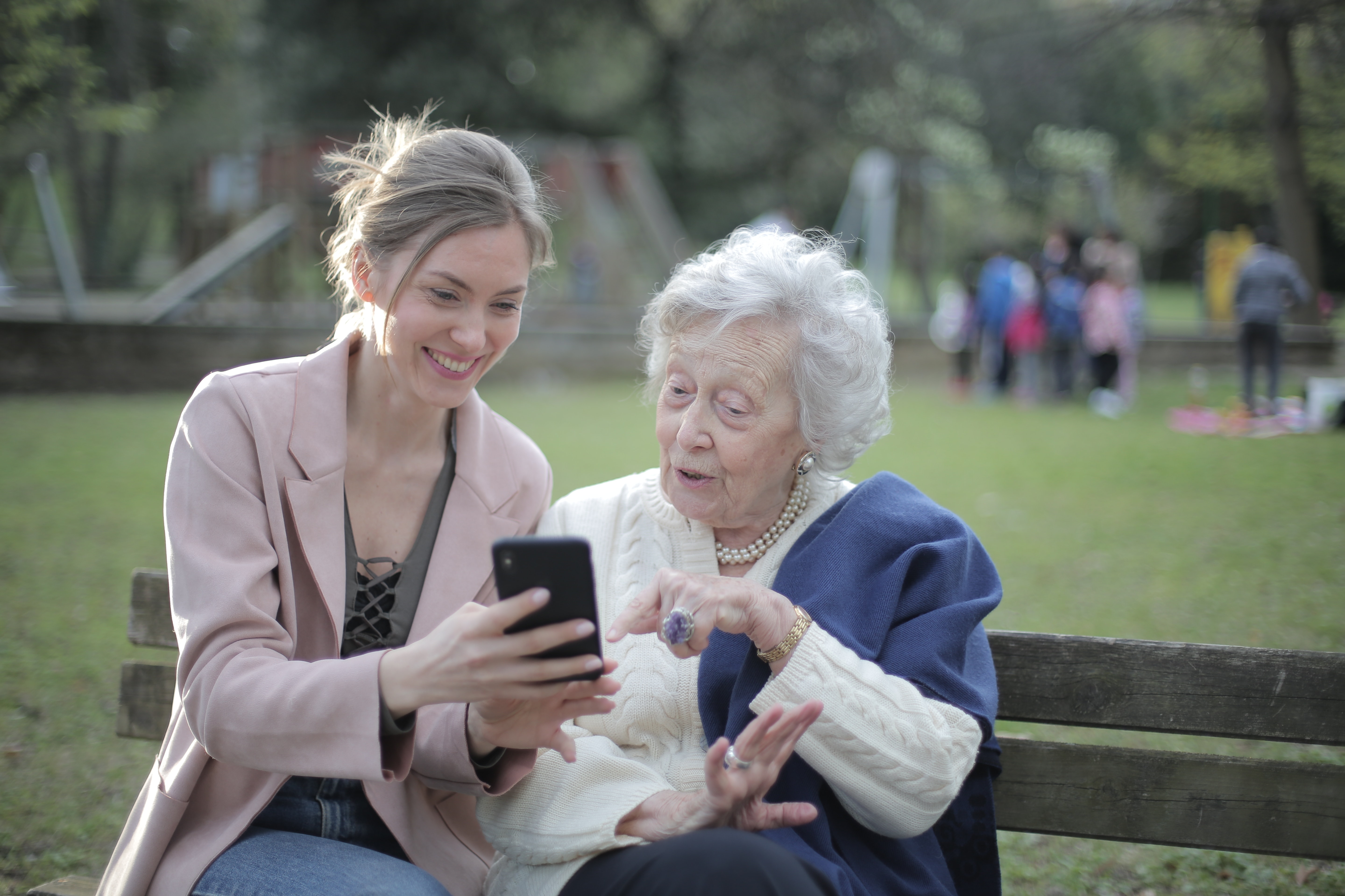 En smilende ung og eldre kvinner sitter på en benk i en park sammen og ser på en telefon