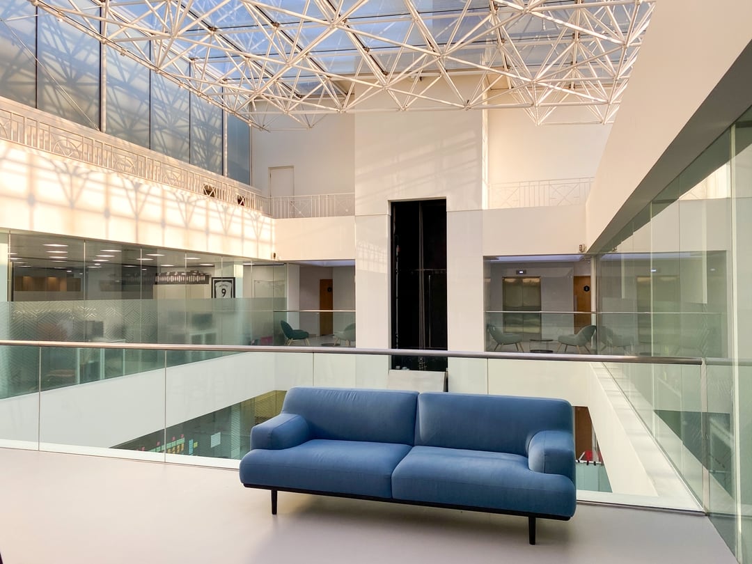 Photograph of the top floor of an open plan office with a large glass ceiling. Blue two seater fabric sofa in the foreground. Behind the chair in the background is an elevator with two blue arm chairs either side.