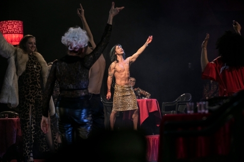 Eugene the Poogene (Orestes), a shirtless Asian man wearing an animal print sarong, illuminated in the background, other performers in the foreground