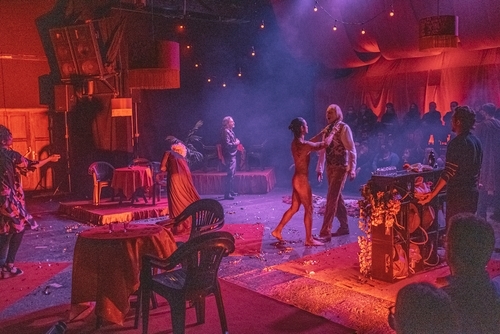 Performers on stage in red lighting, Eugene the Poogene (Orestes), an Asian man, is naked and confronting Svetozar Cvetković (Aegisthus), a white man