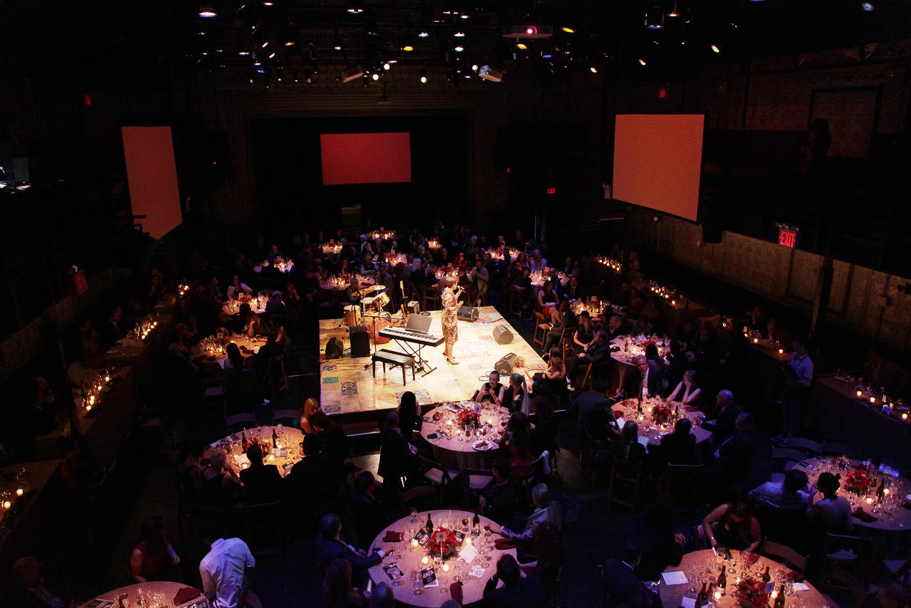 A large dimly-lit venue space with several people sitting around round tables and a raised platform in the center with a performer, piano, drums, and other instruments