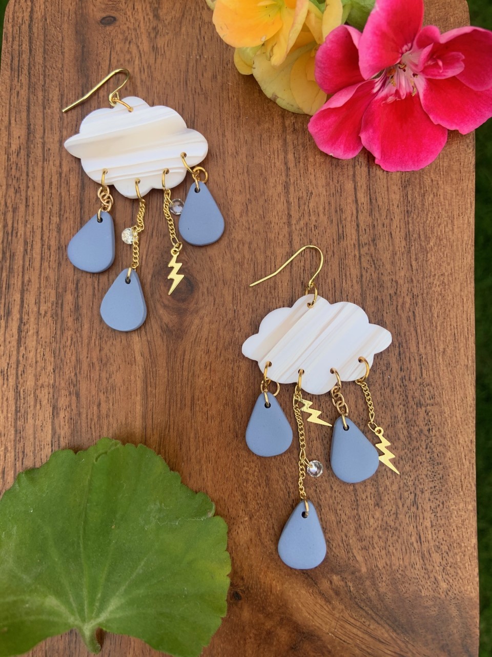Thunderstorm themed polymer clay earrings with a pearl and translucent cloud, accompanied by thin gold lightning bolt chain that dangles three blue rain drops from the cloud.