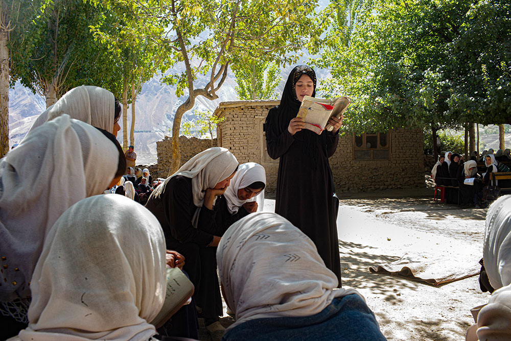 A women wearing black hijab and black clothes standing and reading a book in a group of sitting people