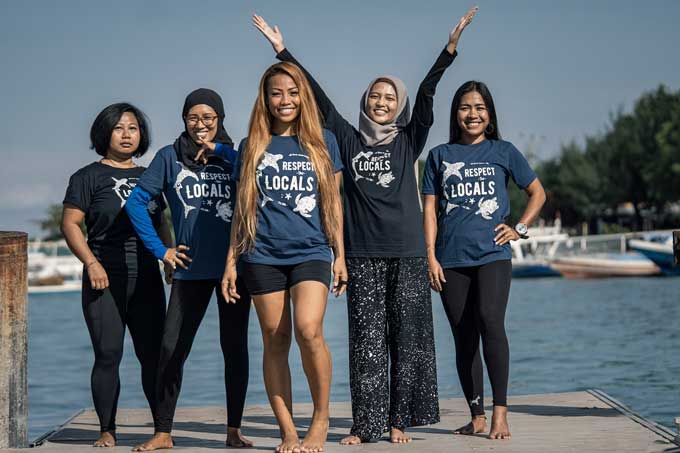 5 women stand near the water, with smiles - wearing shirts showcasing sea creatures and the words Respect Locals