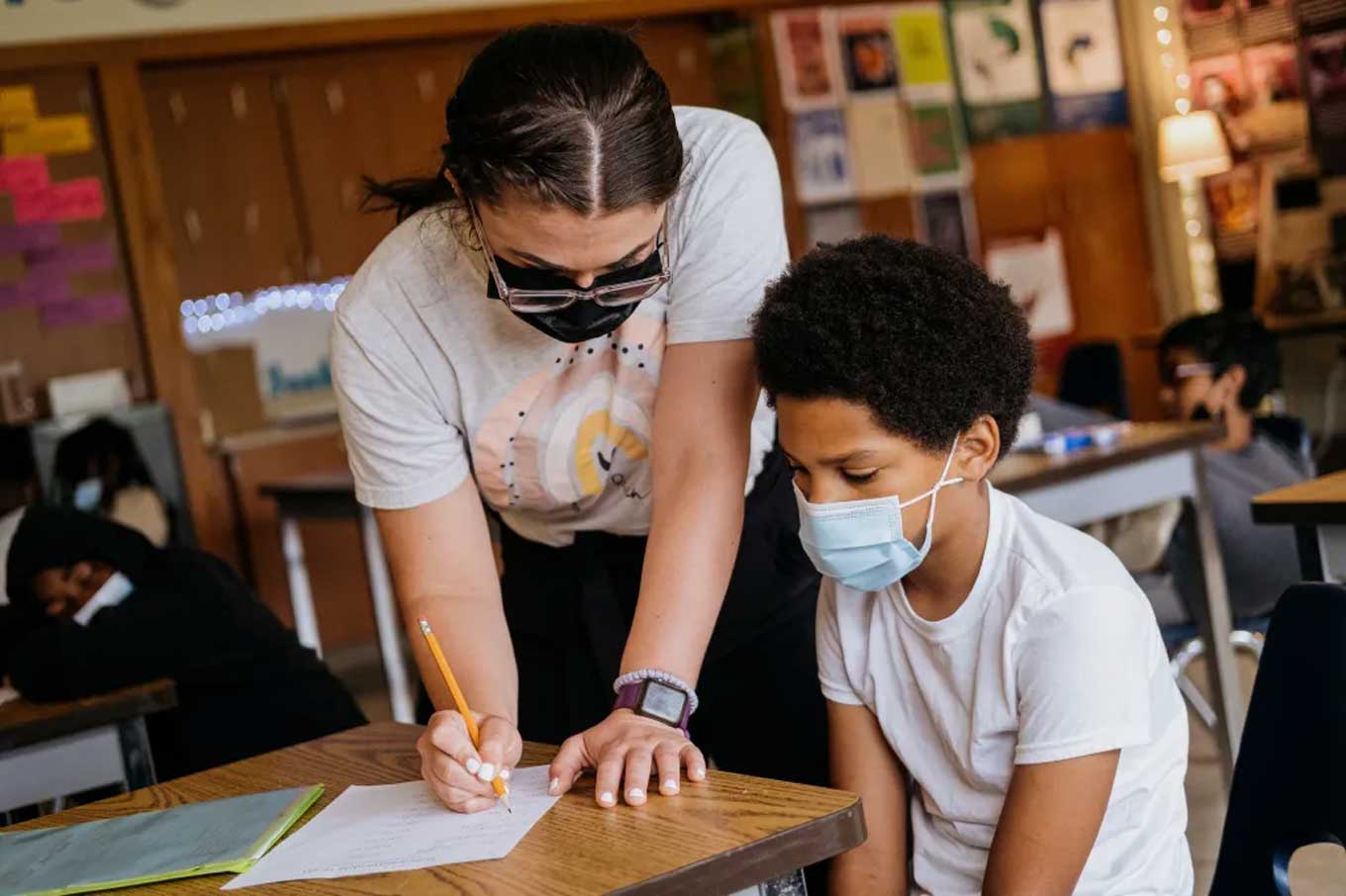A teacher helps a student while both wear face masks