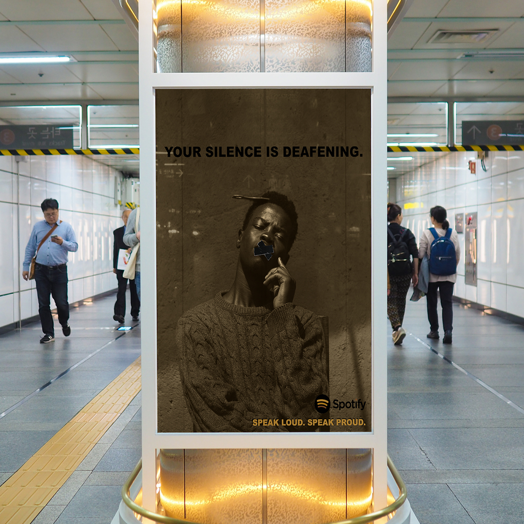 A transit ad in an underground subway passage. The ad is a duotone in black and yellow of an African American with tape over his mouth. His eyes are closed. At bottome right the text reads "Spotify" "Speak Loud. Speak Proud."
