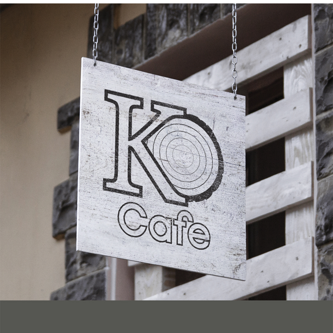 A white and black logo hangs on wooden storefront sign the logo reads "KO café" in black cut out letters. The "O" is drawn like a cross section of a tree trunk.