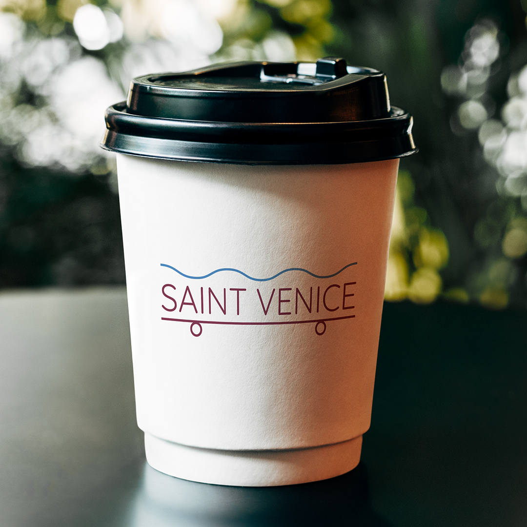 A photo of a white coffee cup with a black lid set on a dark table with greenery blurred in the background. The logo on the cup is set in sans serif dark red font "Saint Venice" there is a line represing a wave pattern above the type. Below a graphic that represents a scateboard.