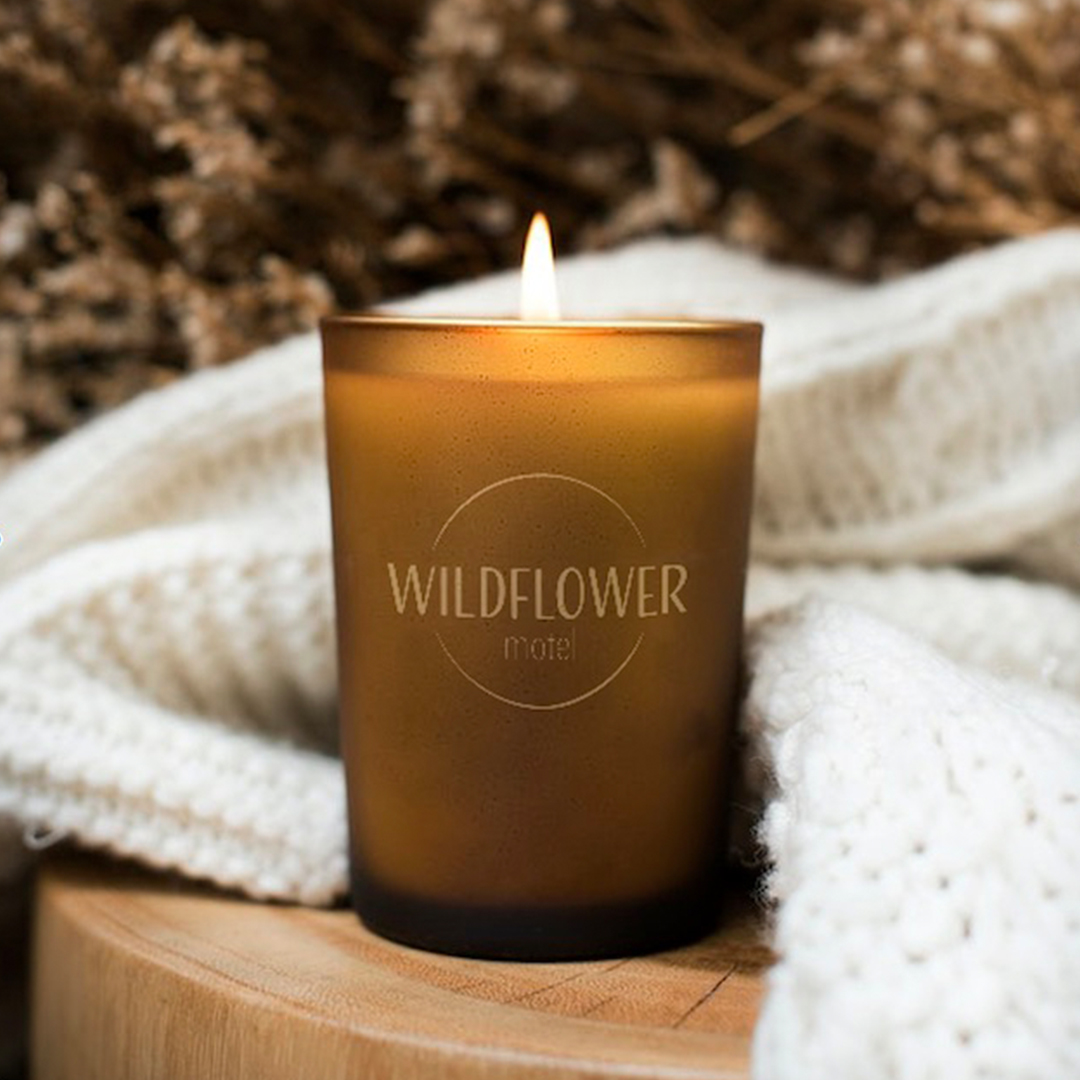 A photo of the Wildflower Motel logo on a image of a lit brown candle. The logo is gold and set within a circle with the text "motel" set below. The font is sans serif.