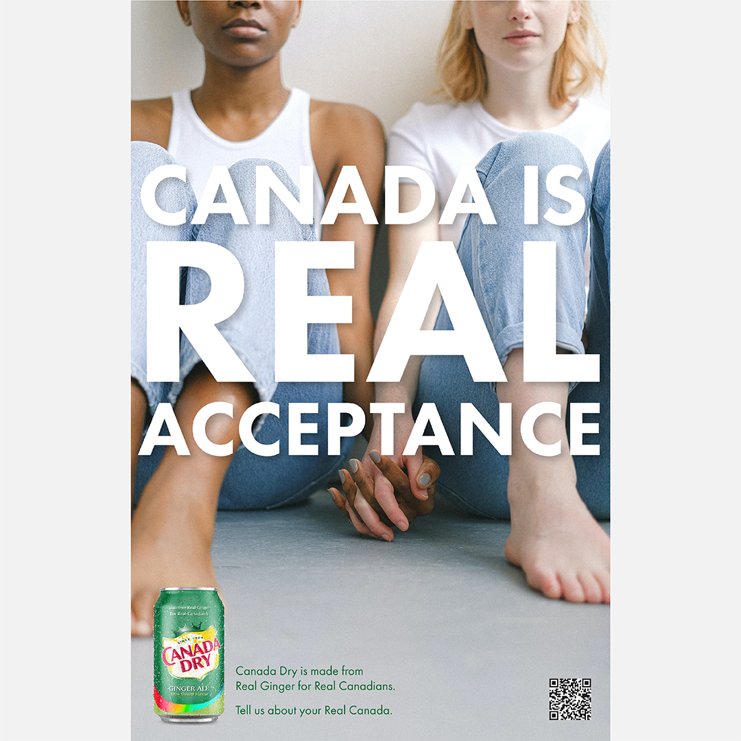 As Canada Dry Gingerale advertisement showing two women seated, wearing jeans and holding hands. One is of African American decent and one is white. The text that appears in the front of the couple in large white letters reads "Canada is Real Acceptance." Below beside the product shot reads "Canada Dry is made from real ginger."