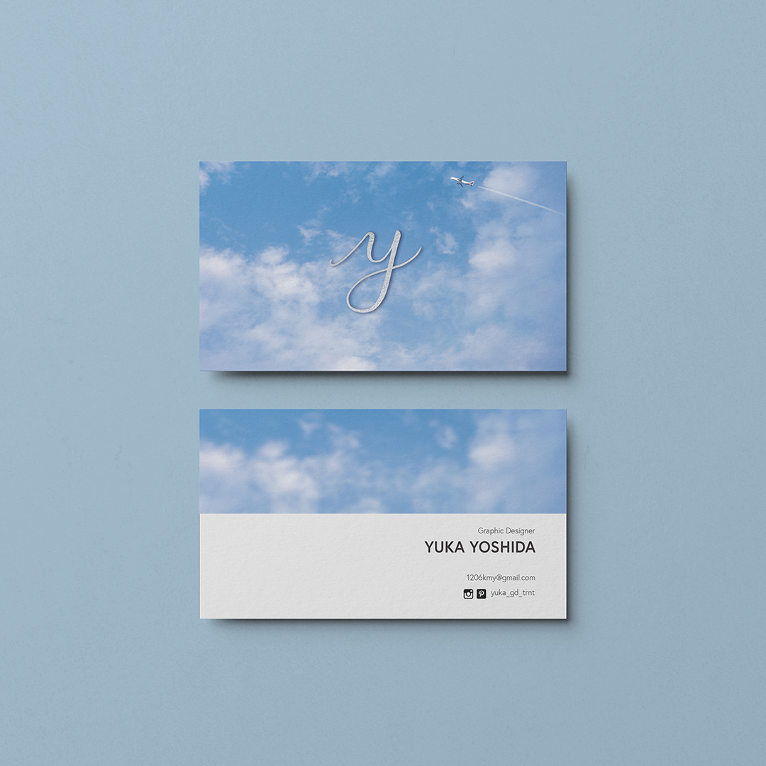 A photo of a business card front and back on a blue background. The card front is a blue sky with a y in script letter. There is a small airplane to the upper right corner. The back of the card is the sky again with the artist's name and title. Graphic design Yuka Yoshida.