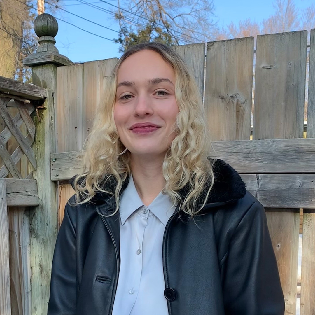 Female identifying person wearing blue shirt and black jacket, smiling, with fence in background