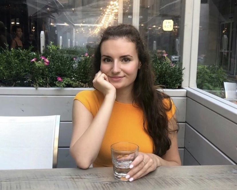 Female identifying person wearing orange top smiling, with hand on face, at an outdoor patio