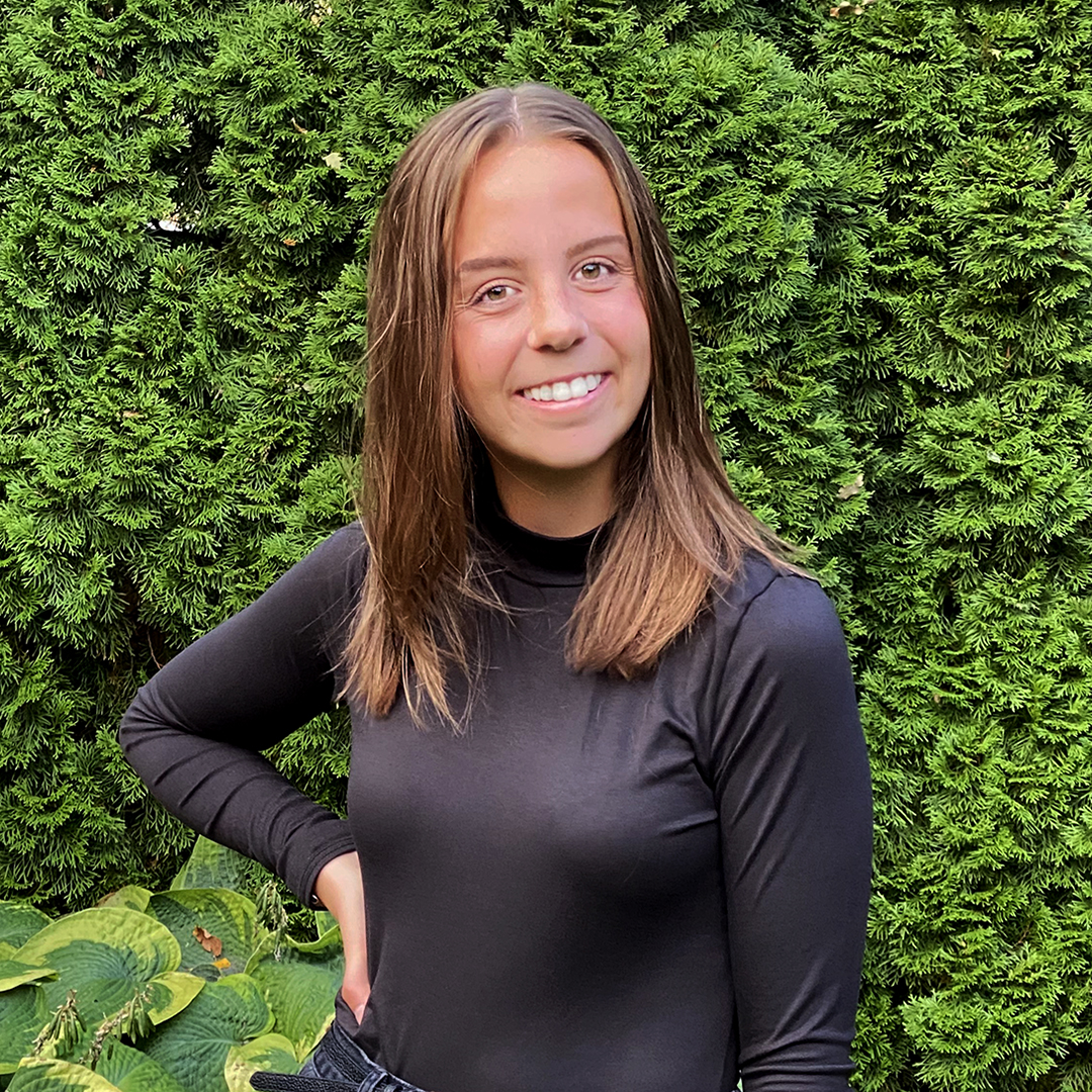 Female identifying person wearing a black top, smiling outside, in front of trees