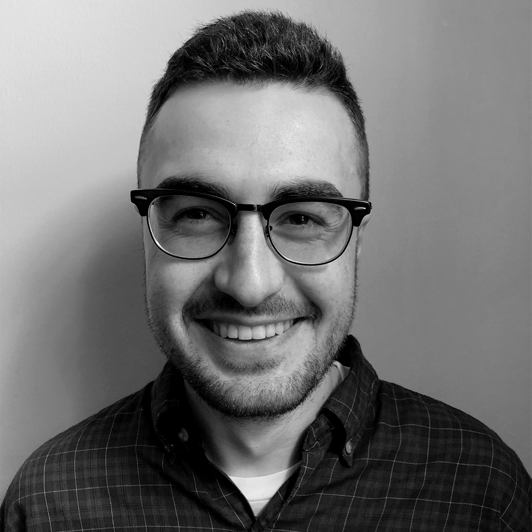 Male identifying person wearing glasses, smiling against grey background