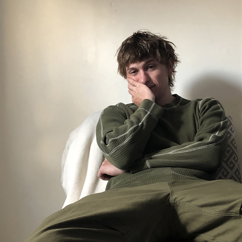 Male identifying person wearing a green shirt sitting in chair, with hand on face.