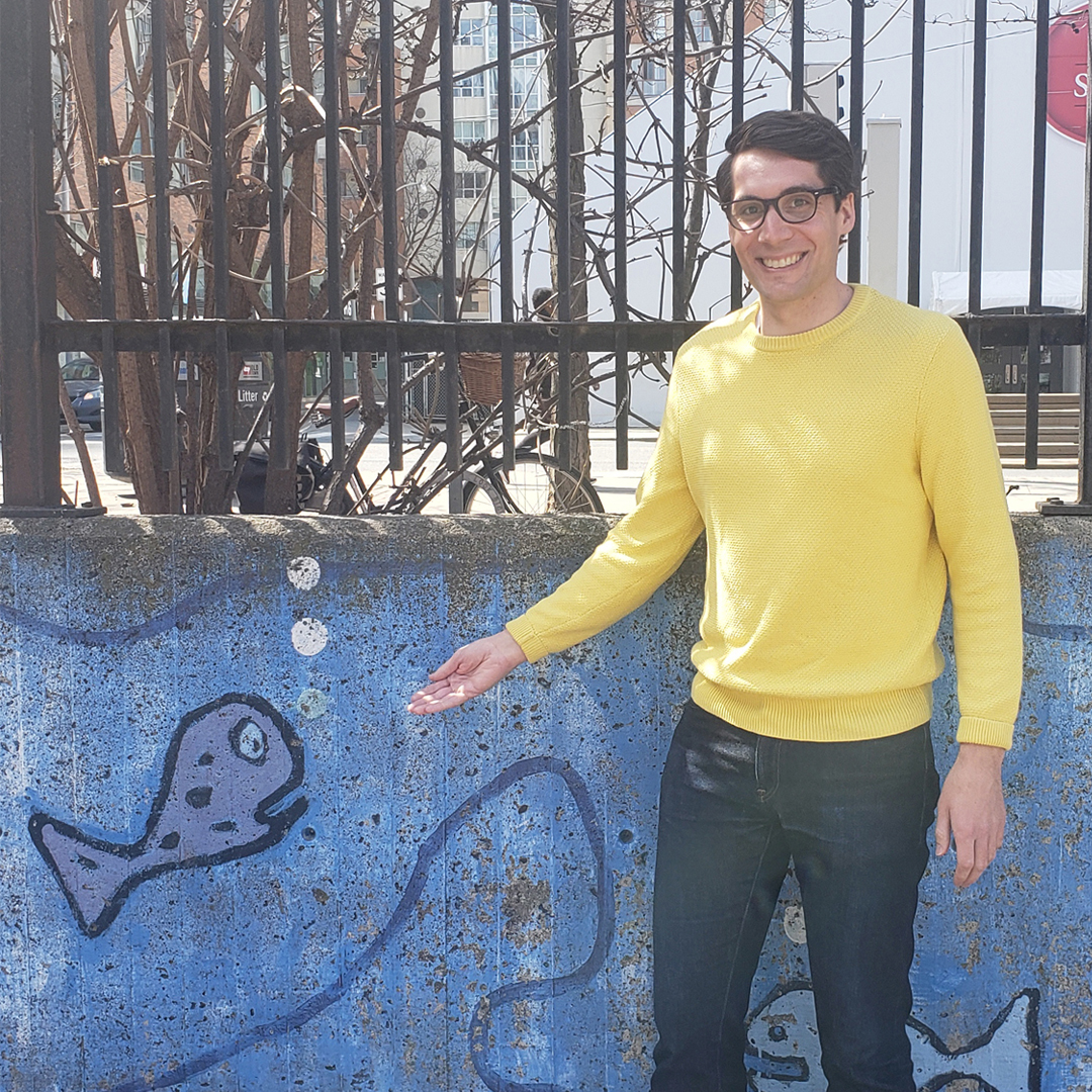 Male identifying person wearing glasses and a yellow shirt, gestures at fish painting, outside