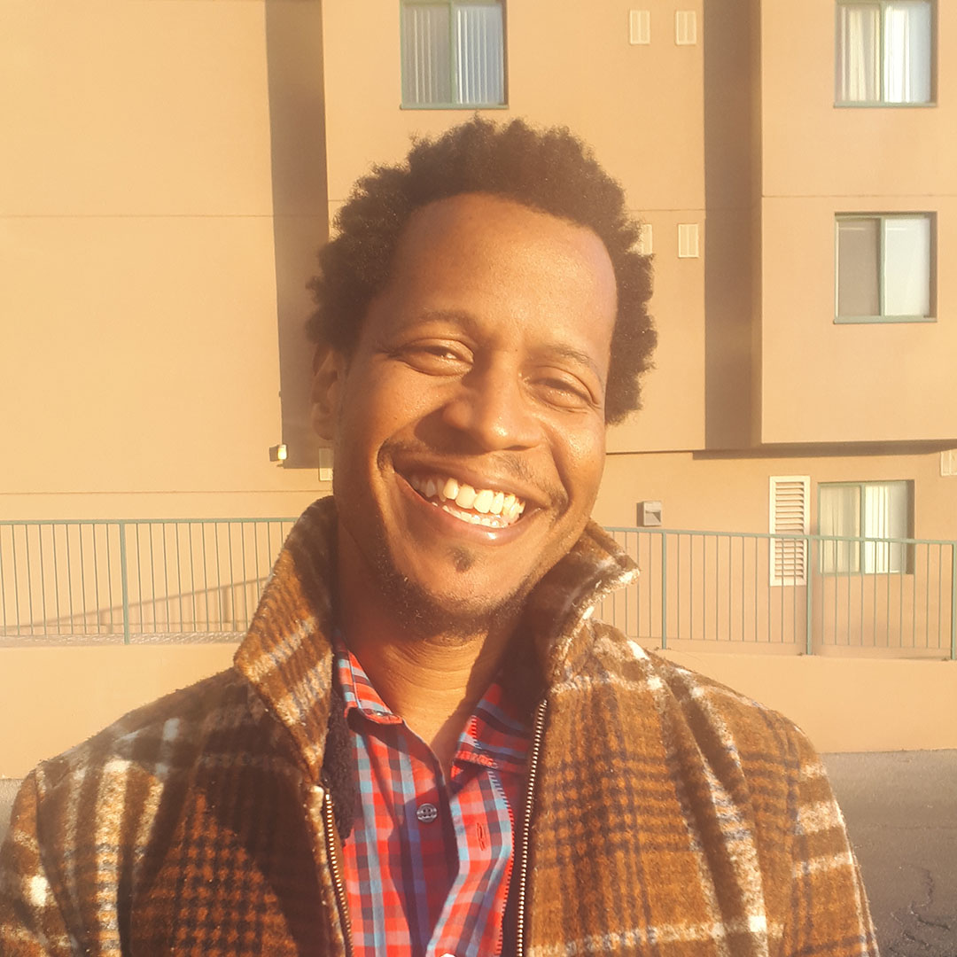 Male identifying person wearing a checked coat, smiling outside, in front of a building