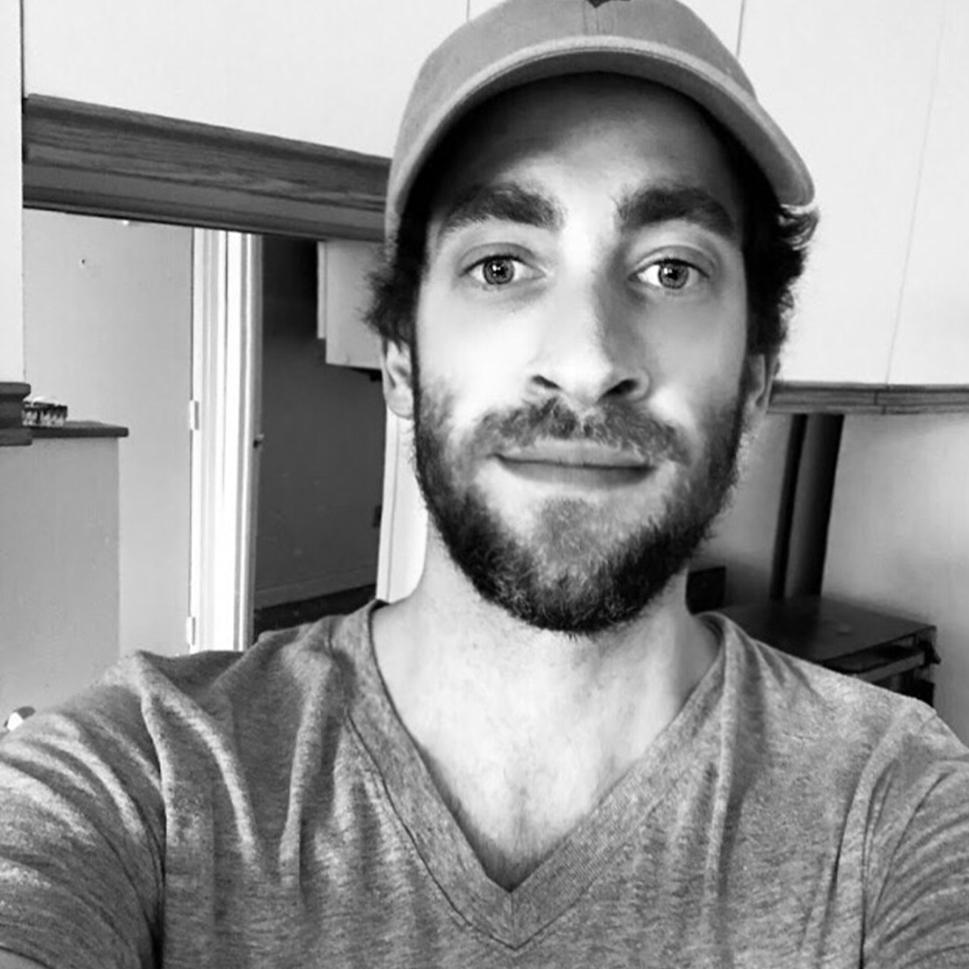 Male identifying person with beard, wearing ball cap, smiling, inside