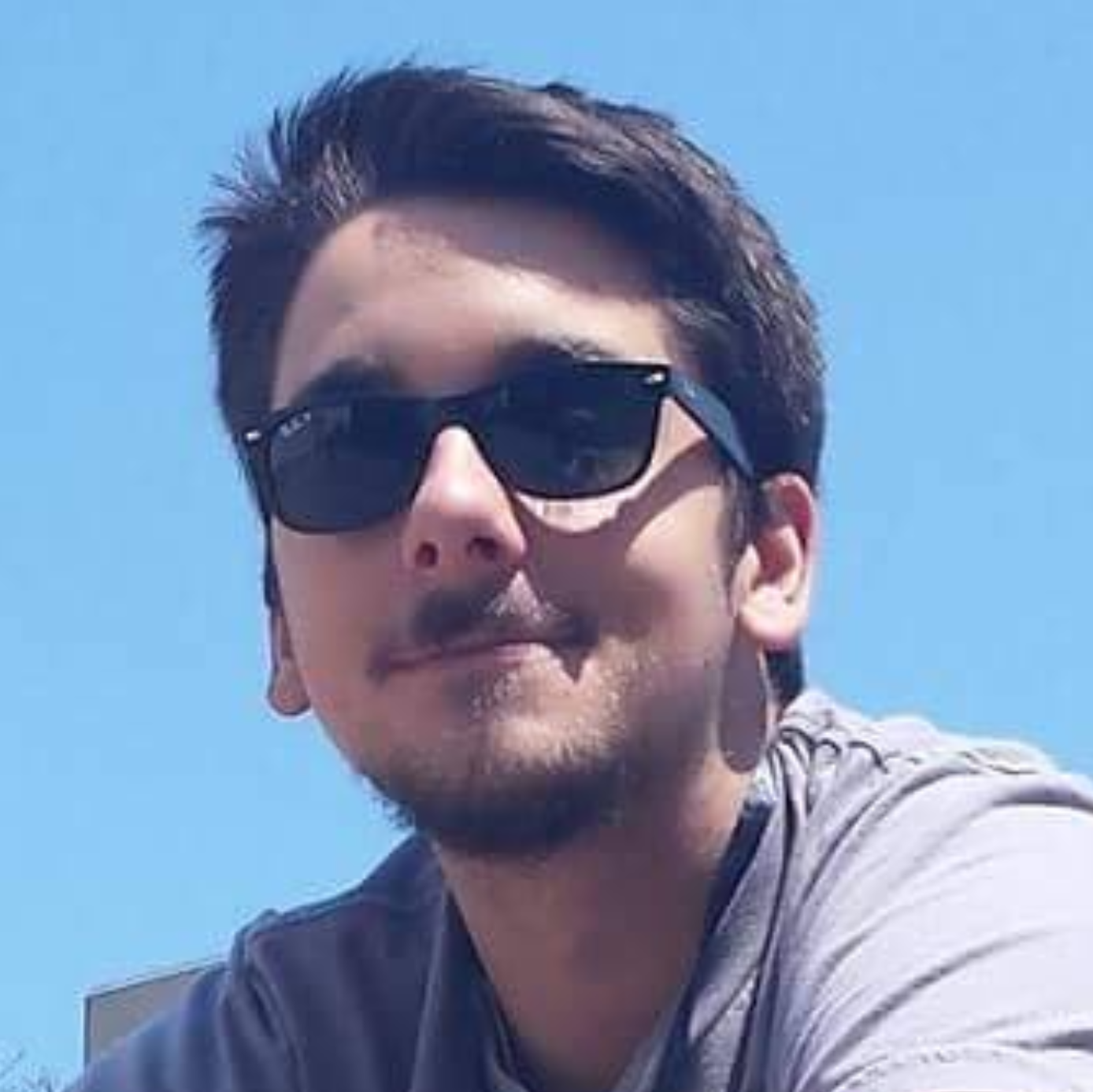 Male identifying person, wearing sunglasses and smiling, against blue sky background