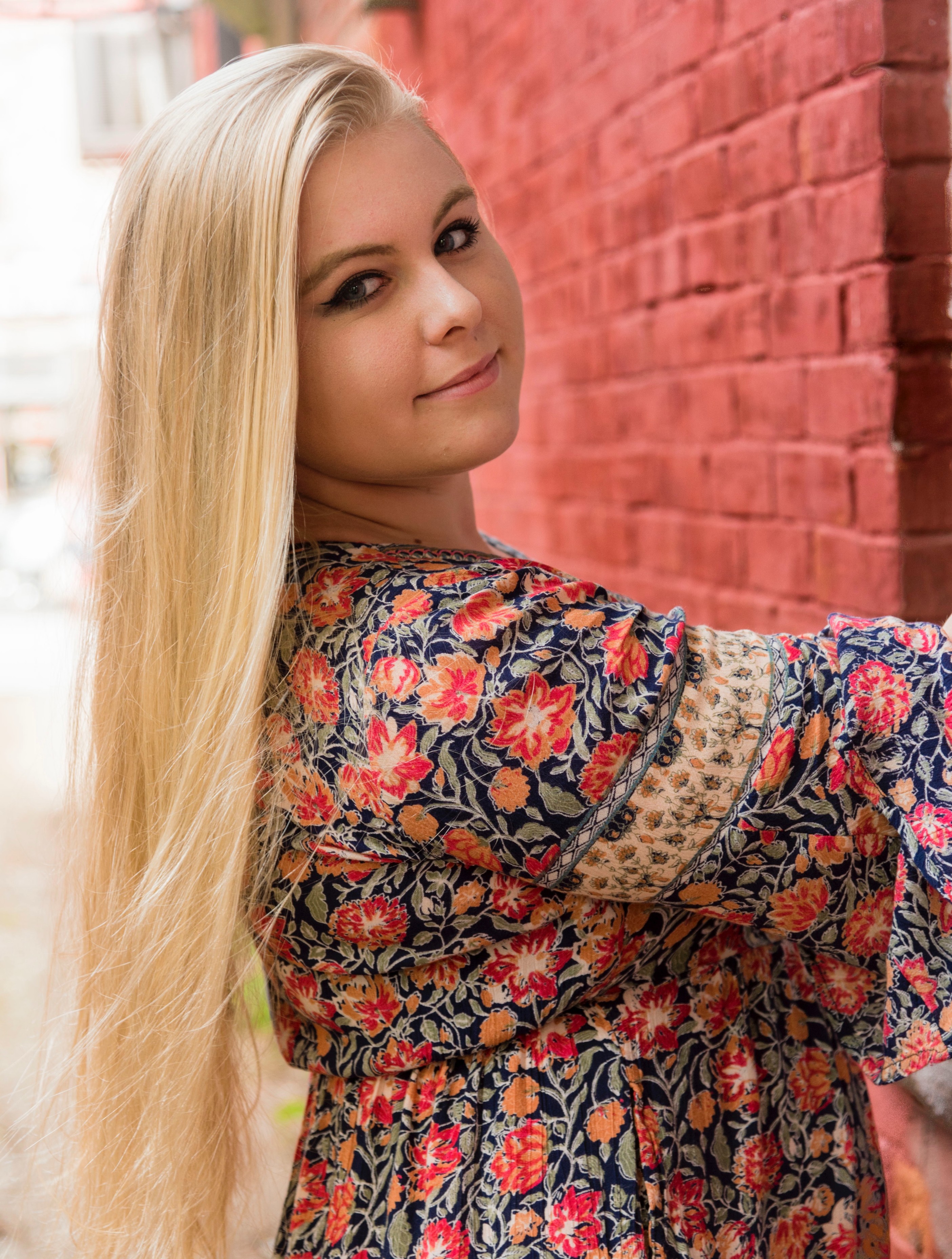 Picture of Molly, outdoors and smilig at the camera, wearing a floral dress navy, red and orange.