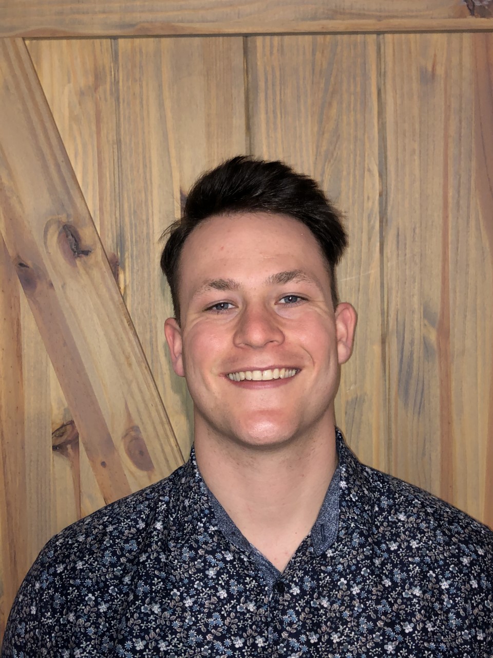 Picture of Riley, posing in from of the camera smiling and wearing a dark blue shirt with light bluse flowers