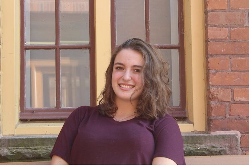 Picture of Shania, posing and smiling outside, in front of a window, wearing a purple shirt.