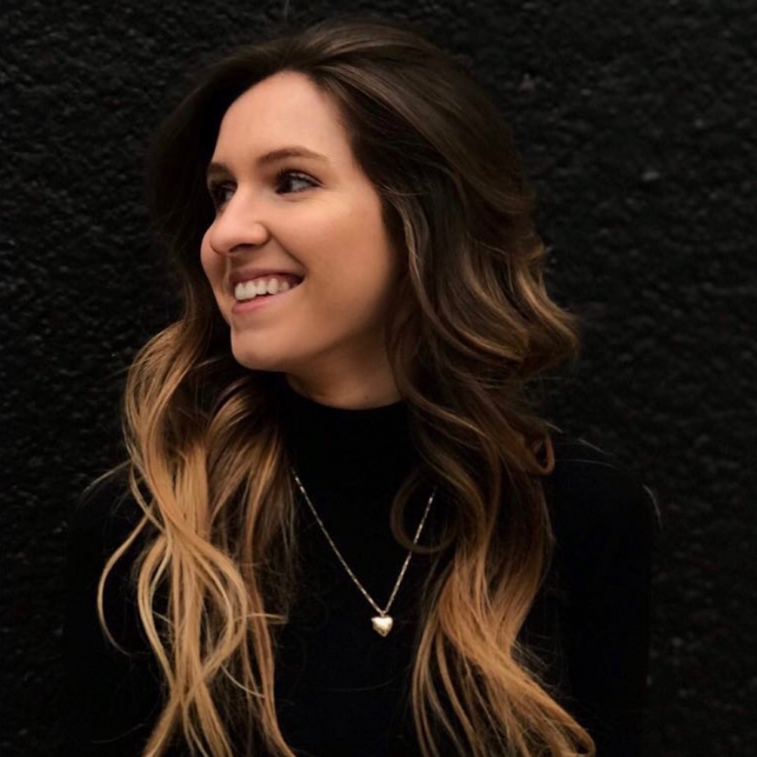 A headshot of Sadie Maycock standing in front of a black wall, smiling.