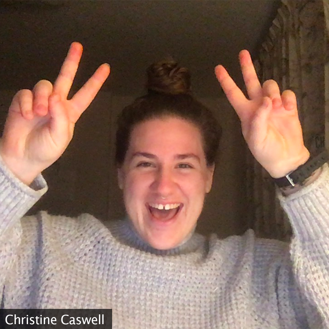 Picture of creator in a light grey turtleneck sweater smiling at the camera and she is throwing up a peace sign on both hands at each side of her.