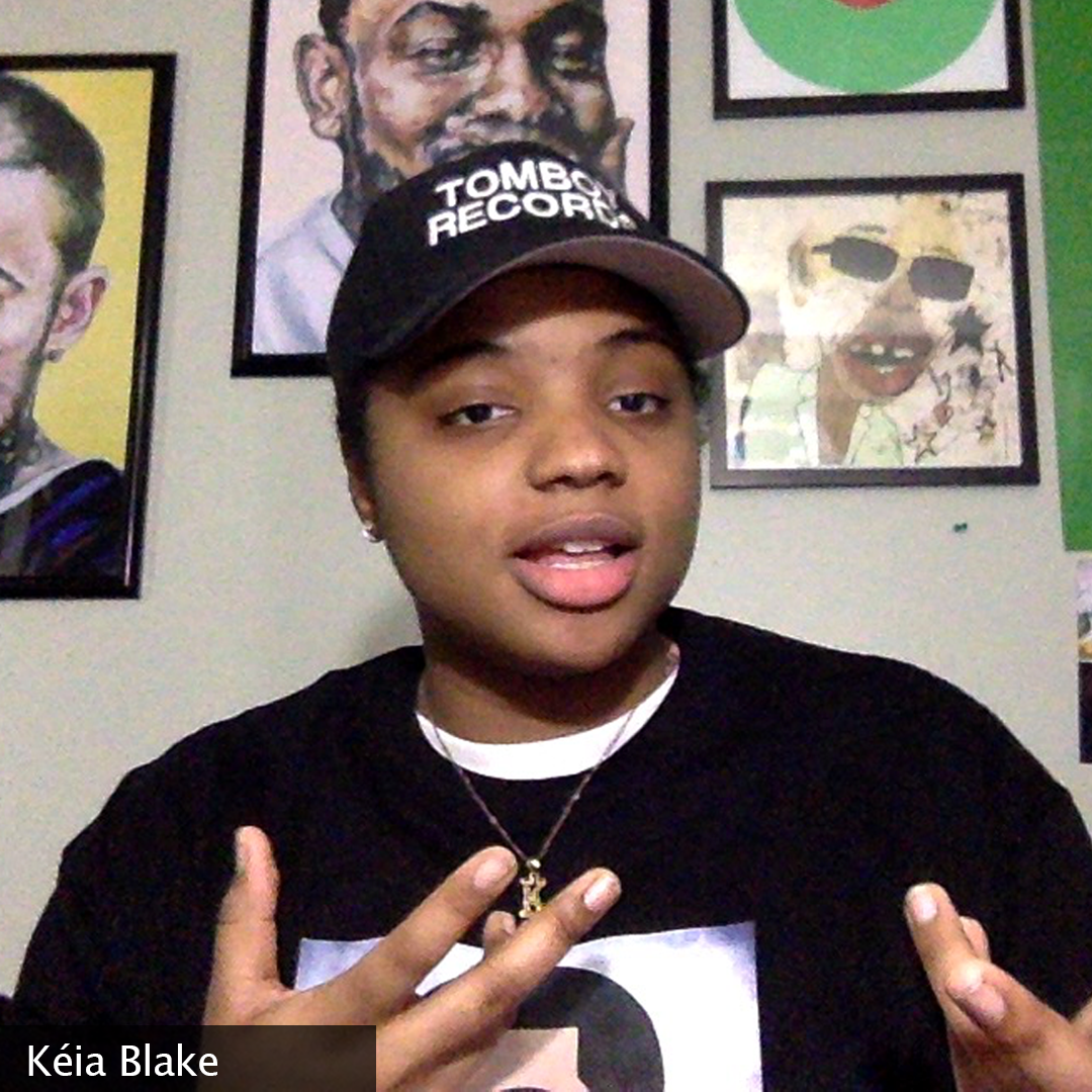 Photo of Keia mid statement wearing a “Tomboy Records” hat. The wall behind her has posters of several hip hop artists:
