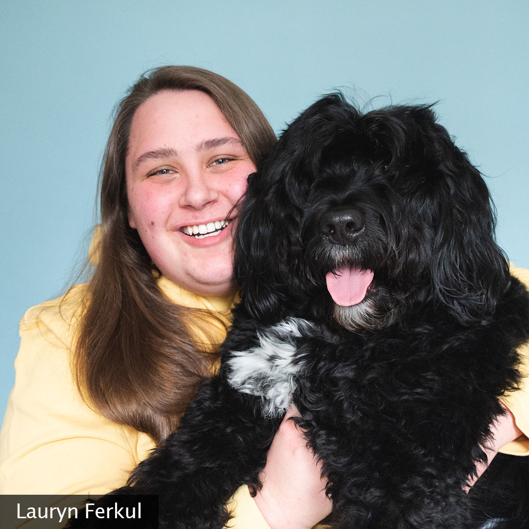 Lauryn is smiling at the camera wearing a yellow hooded sweatshirt and her black, fluffy dog, Olie, is snuggled beside her.