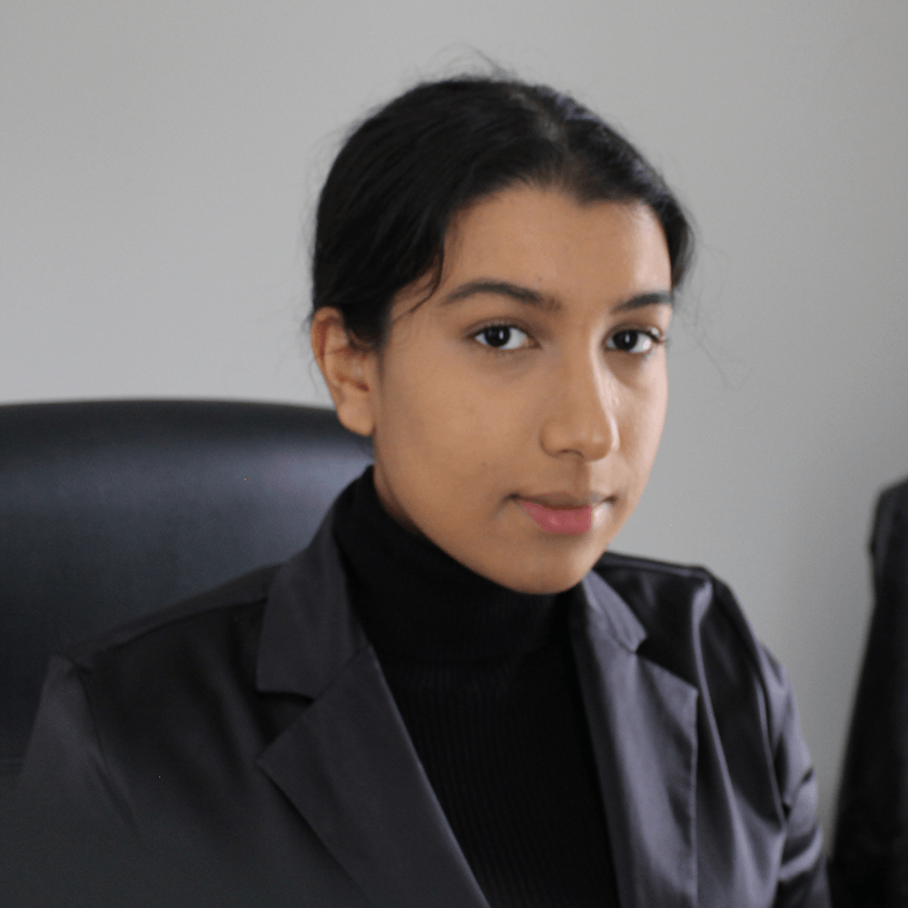 Aisha sitting and looking at the camera wearing a black turtle neck t-shirt with a light-black blazer