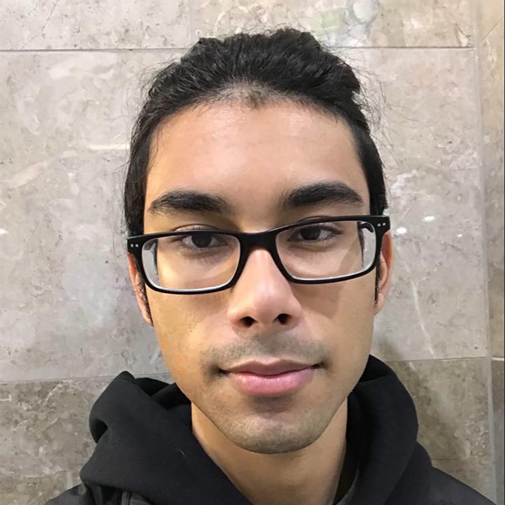 Picture of Alex Sumeer against a wall, wearing glasses and a black hoodie