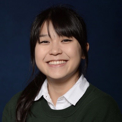 Picture of Maya, smiling to the camera and wearing a black sweater with a white shirt
