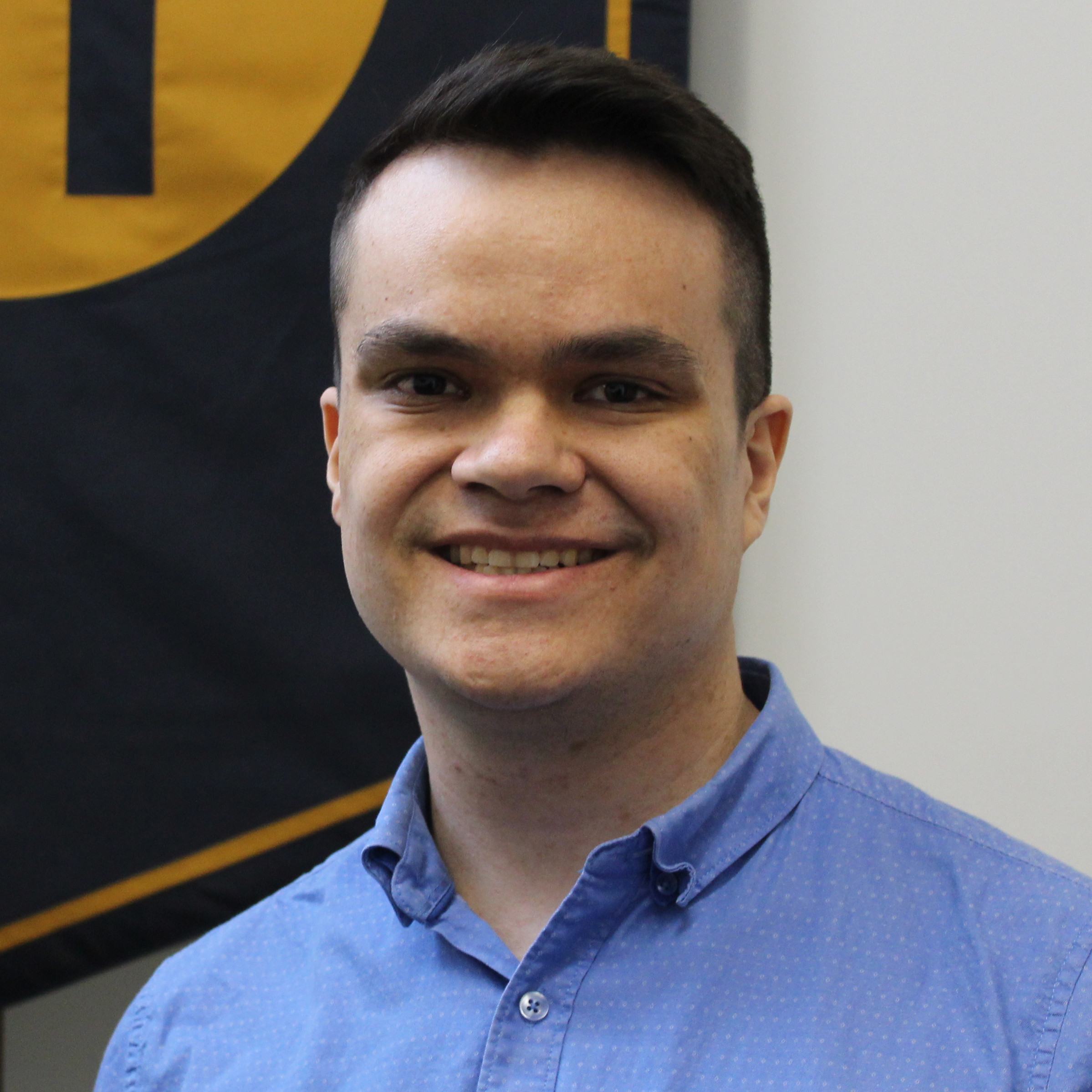 A headshot of Joshua Wood. They are wearing a blue button-up shirt. They are in front of a school banner which is hanging on the wall behind them.