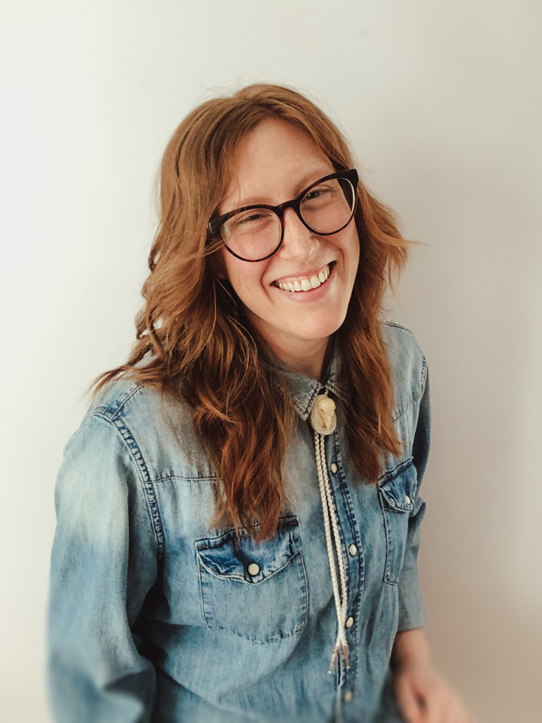 A headshot of Sandra Mannila. They are sitting in front of a pale grey wall, and are wearing a denim button-up shirt and a white leather bolo tie.