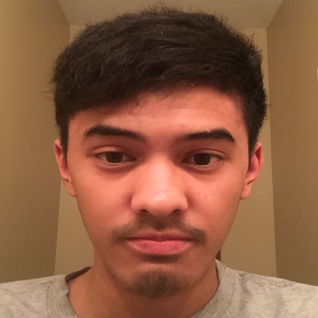 Daniel Regalado, 20 year old, Filipino male with short black hair and a stubble Balbo. Wearing grey T-shirt in front of warm lighting.