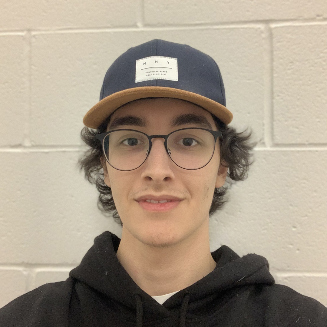 Lazar, a younger white man smiles at the camera. He is wearing glasses, and a baseball hat with curly brown hair protruding from beneath it.