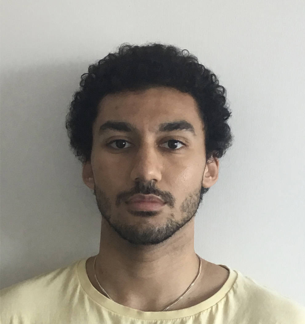 Tall brown male with curly hair, a beard, wearing a yellow t shirt standing behind a white door