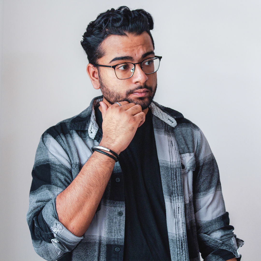 A portrait of Sheel Darbar, a Canadian artist with curly black hair, wearing black-rimmed glasses and a flannel.