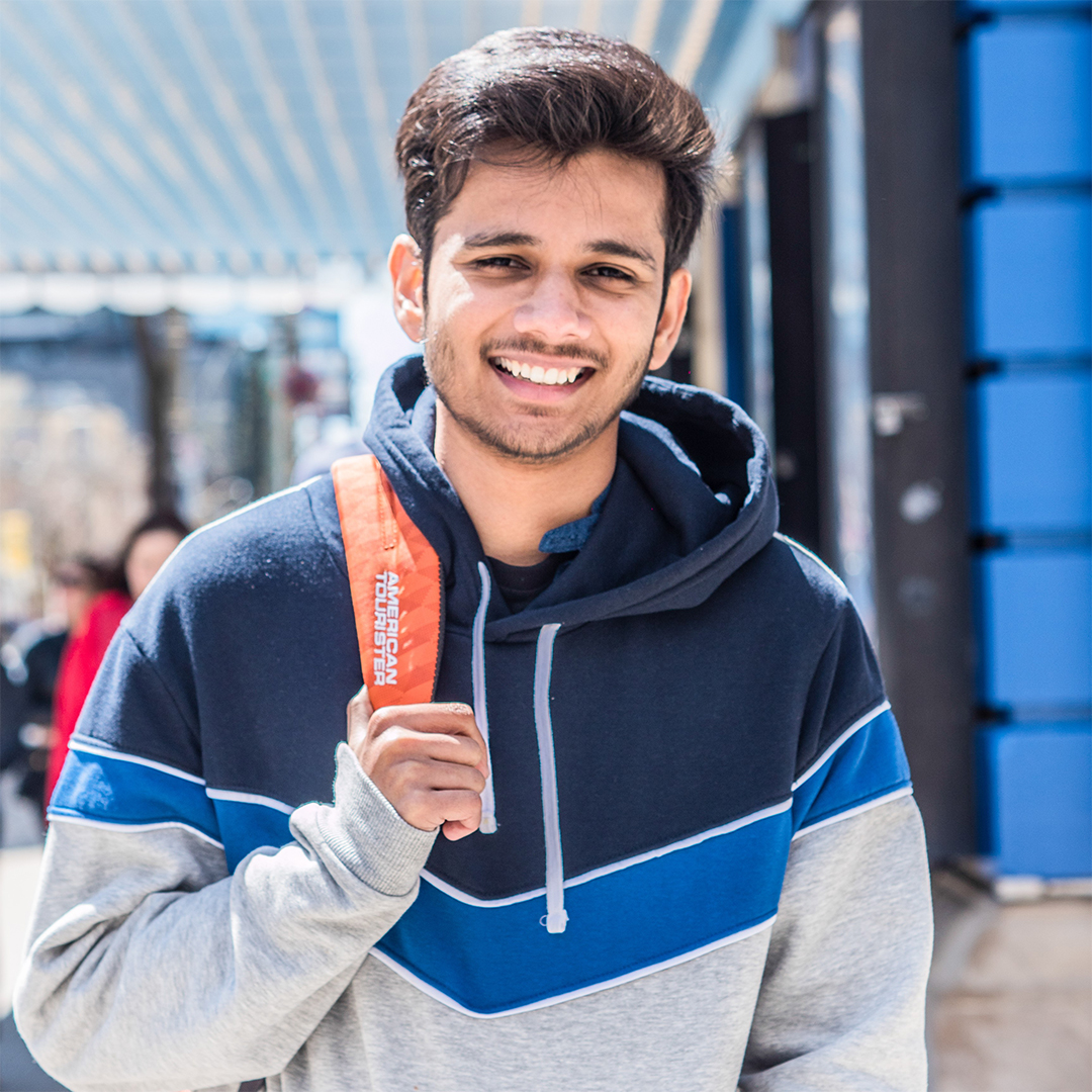 Shubham, an Indian male with black hair, is outside on a bright day. He wears a blue-grey hoodie with an orange backpack and smiles at the camera.