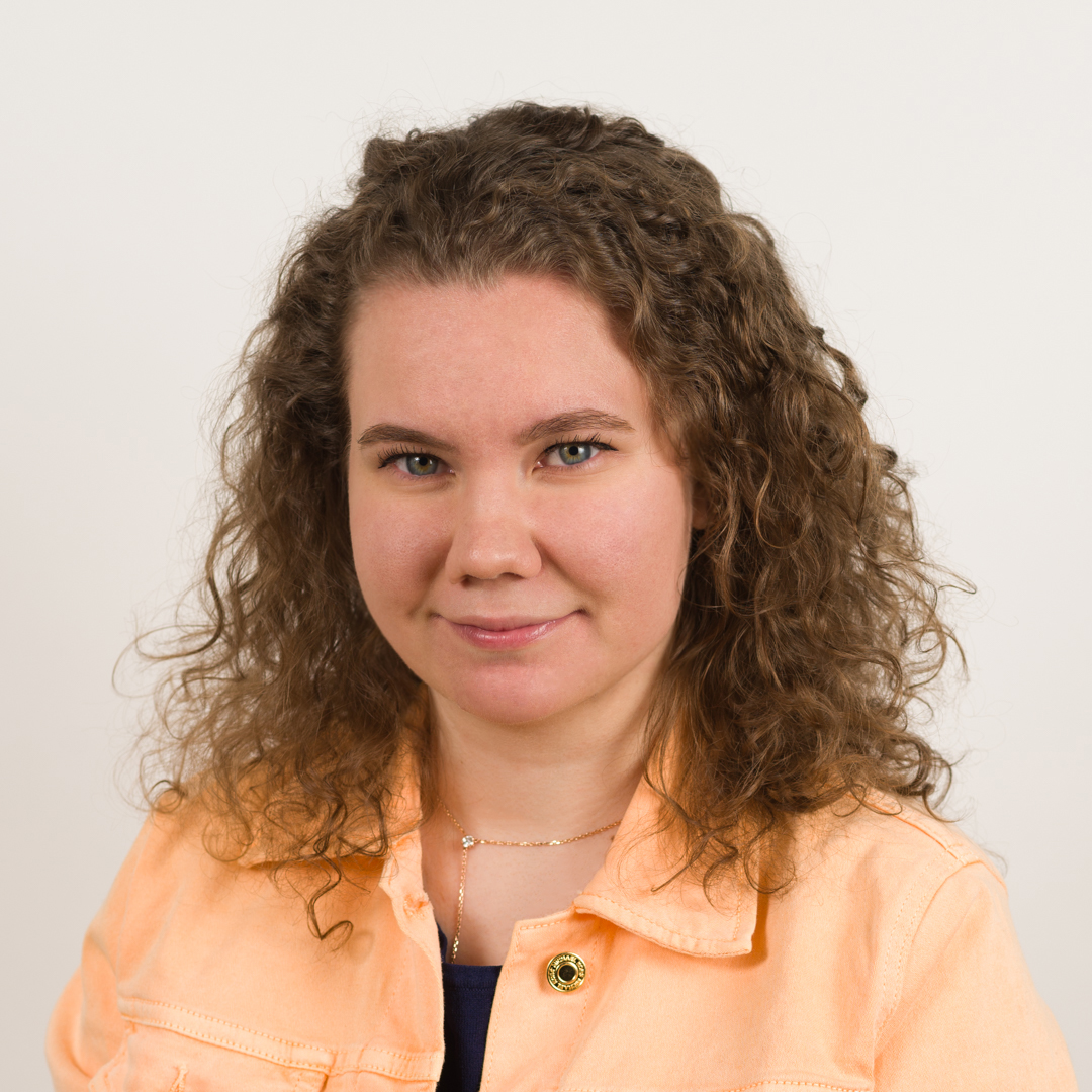 A picture of Viktoriia with curly hair and a smile, wearing a peach-colored jacket on a white background.
