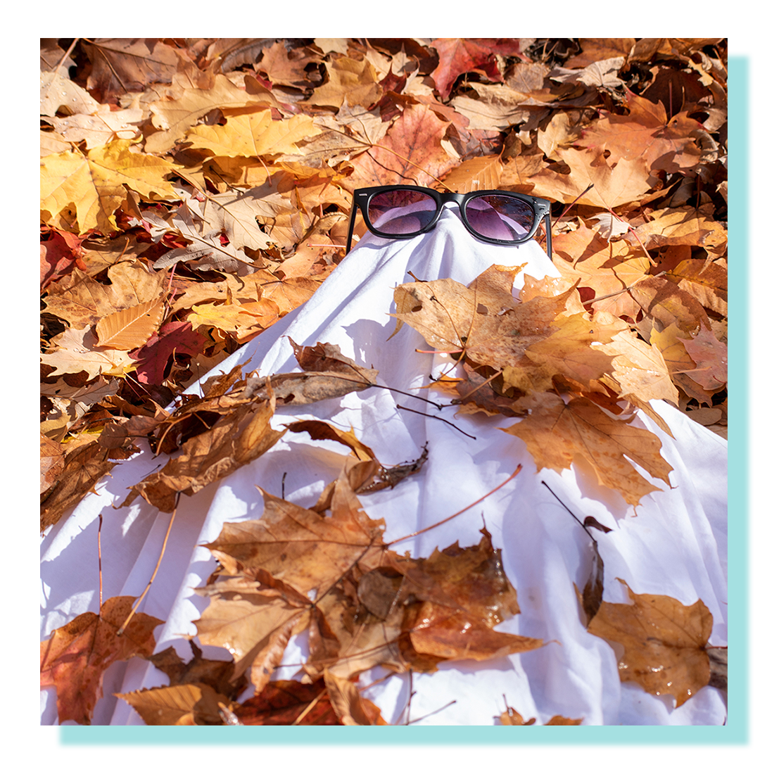 A person in a dressed as a ghost wearing glasses and lying on a bed of leaves.