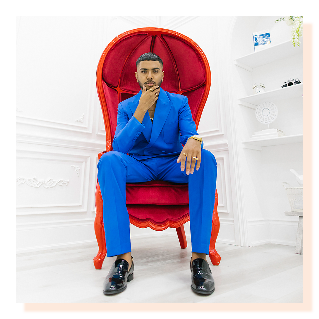 A man wearing a bright blue suit sitting on large royal-looking bright red chair. Man’s face in hands.