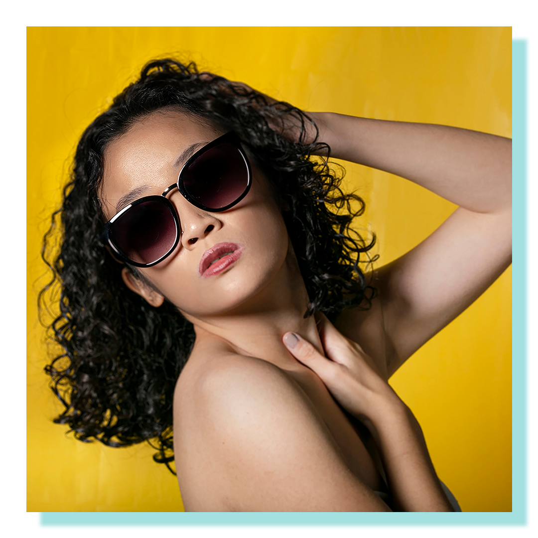 Portrait of a woman waist up, with sunglasses against a vibrant yellow background.