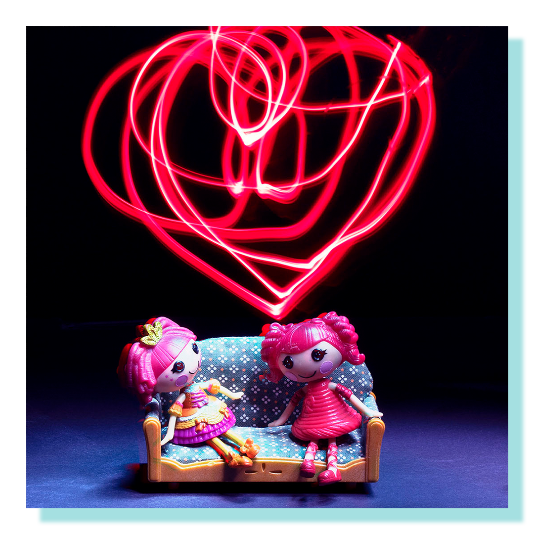 Two small toy dolls sitting on a dollhouse sofa with a red heart light in the background.