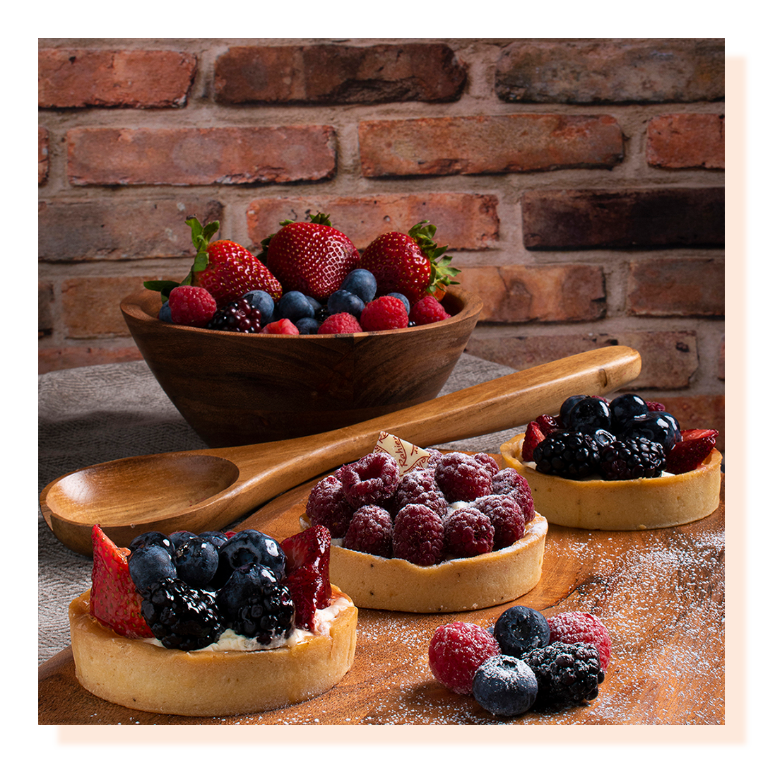Berry tarts and berry bowl of fruit on a wooden chopping board and brick wall in the background.