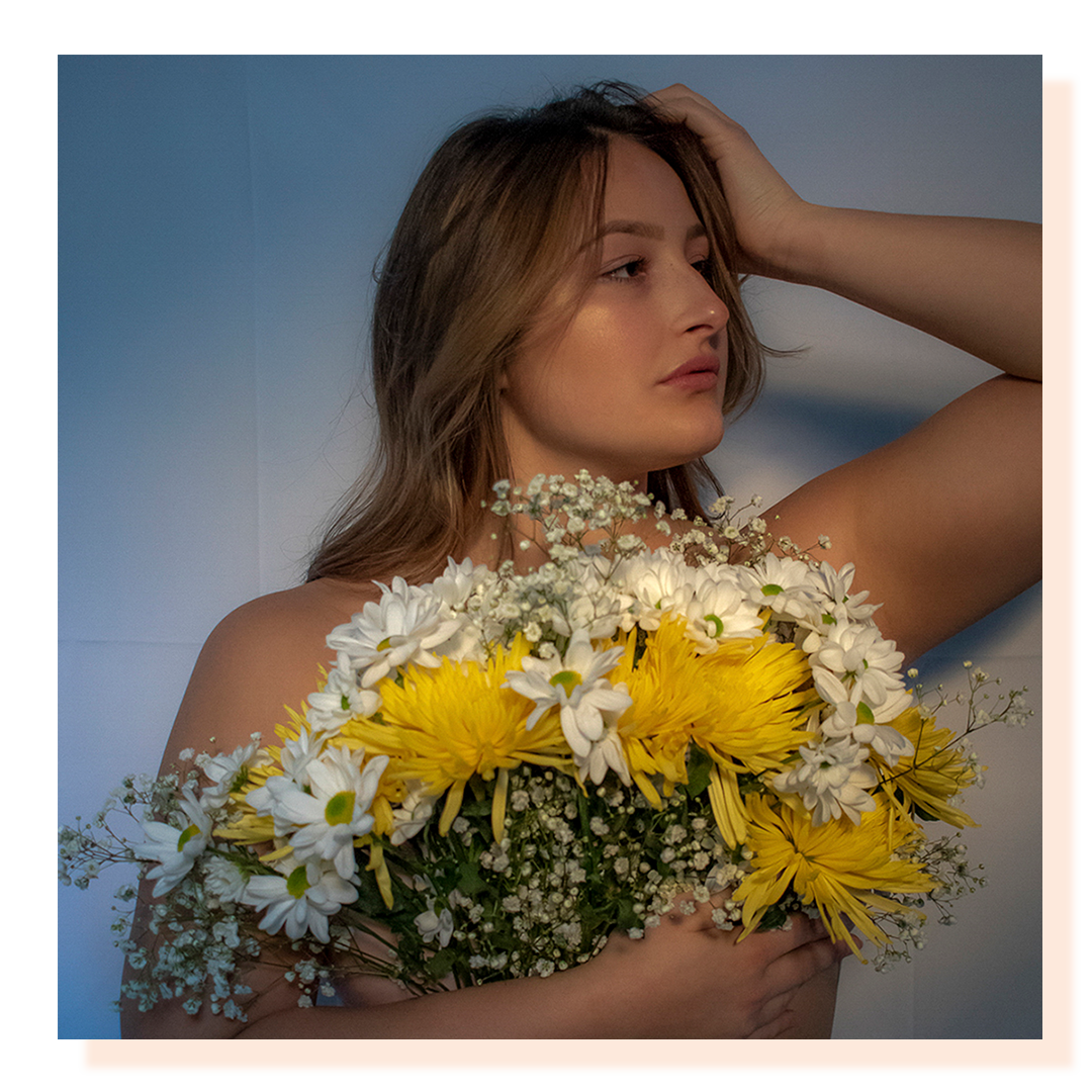 A girl in front of blue background holding a large bouquet of yellow and white flowers in front of her chest.